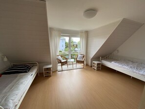 A bedroom with a sloped ceiling, a bed on the left, a chair and table by the window, and a second bed on the right.