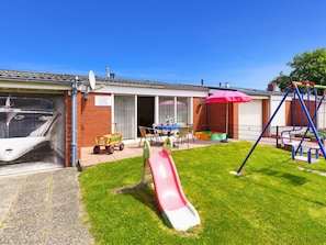 Un jardin à l’arrière d’une maison, aménagé avec un toboggan, une balançoire, une table et des chaises, ainsi qu’une petite piscine.