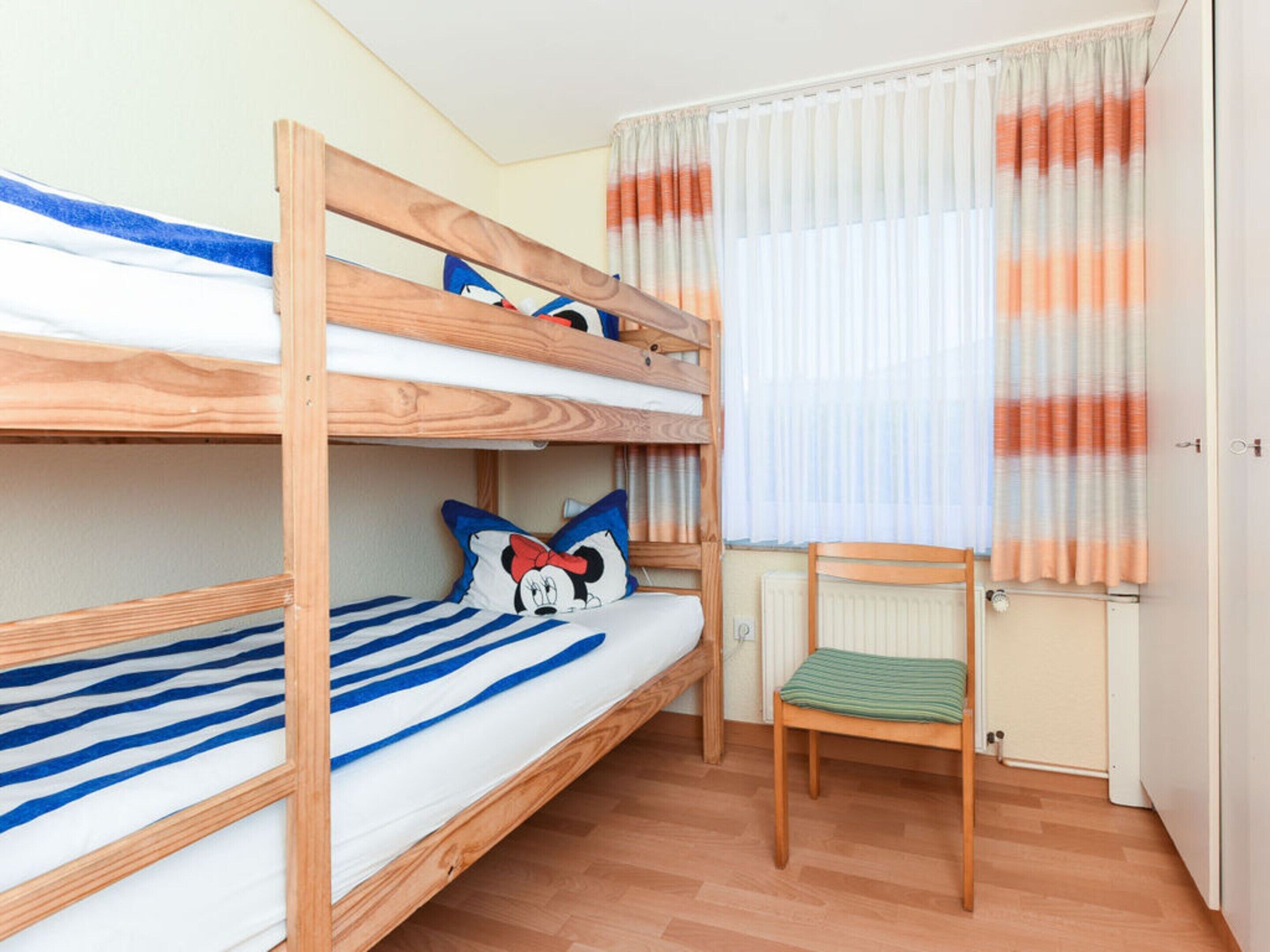 A bunk bed room with a Mickey Mouse pillow, a chair, and a window with curtains.