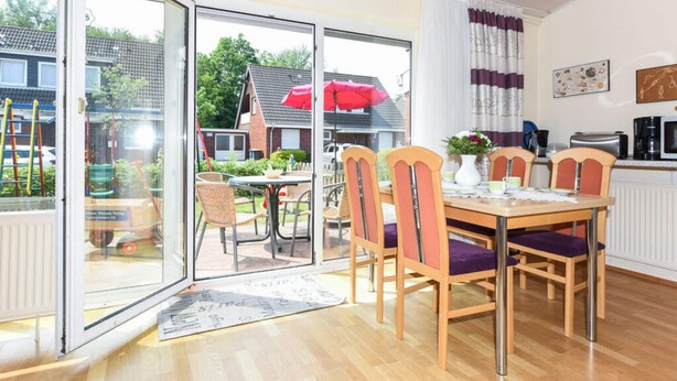 A dining area with a table and chairs, a kitchen area with appliances, and a view of the outside through a glass door. - Holiday Home Weisse Robbe (Esens)