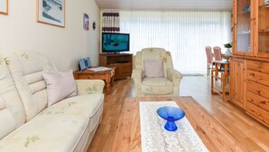 A living room with a sofa, armchair, wooden coffee table, TV, and a dining area with chairs and a cabinet. - Holiday Home Weisse Robbe (Esens)