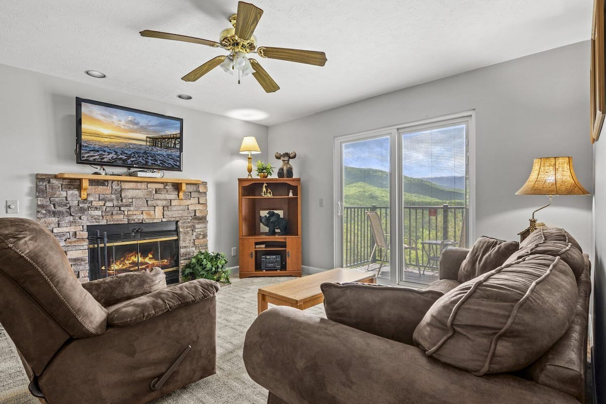Ein Wohnzimmer mit Kamin aus Stein, zwei braunen Sofas, einem Couchtisch, einem Fernseher über dem Kamin, einem Deckenventilator und einem Balkon mit Blick auf Hügel.