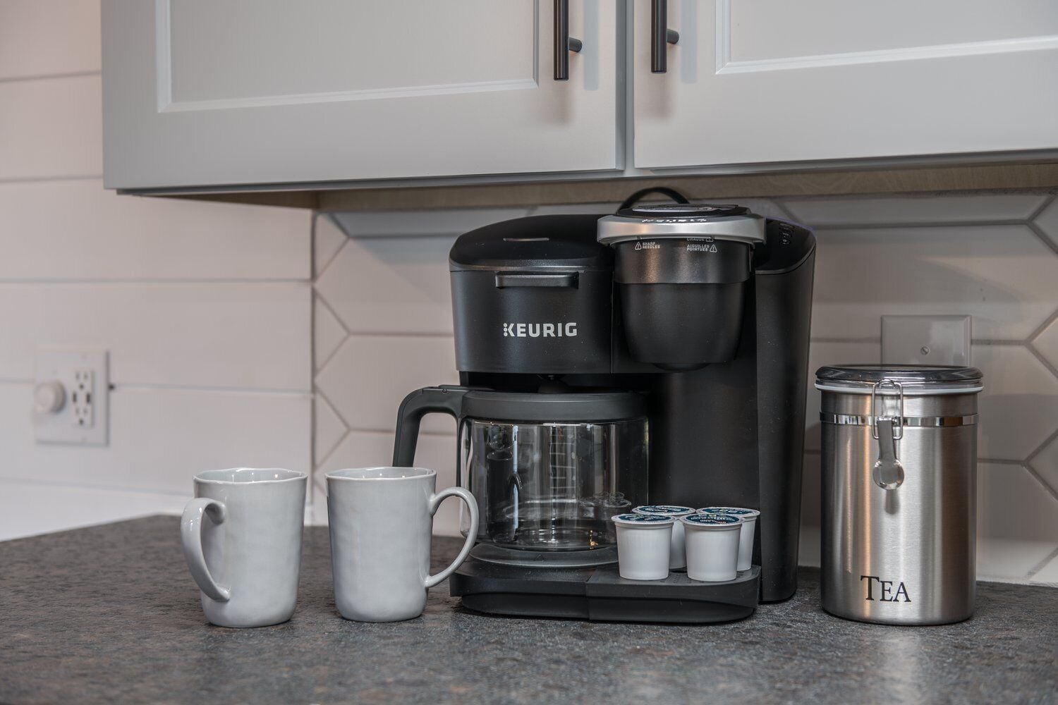 Una cafetera Keurig con una jarra de café, dos tazas y un recipiente de acero inoxidable para té sobre una encimera de cocina.