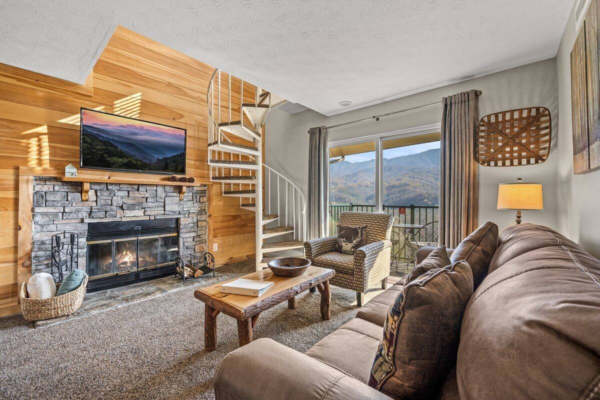 Ein gemütliches Wohnzimmer mit Kamin aus Stein, einer bequemen Couch, einer Holztreppe und einem großen Fenster mit Blick auf die Berge.