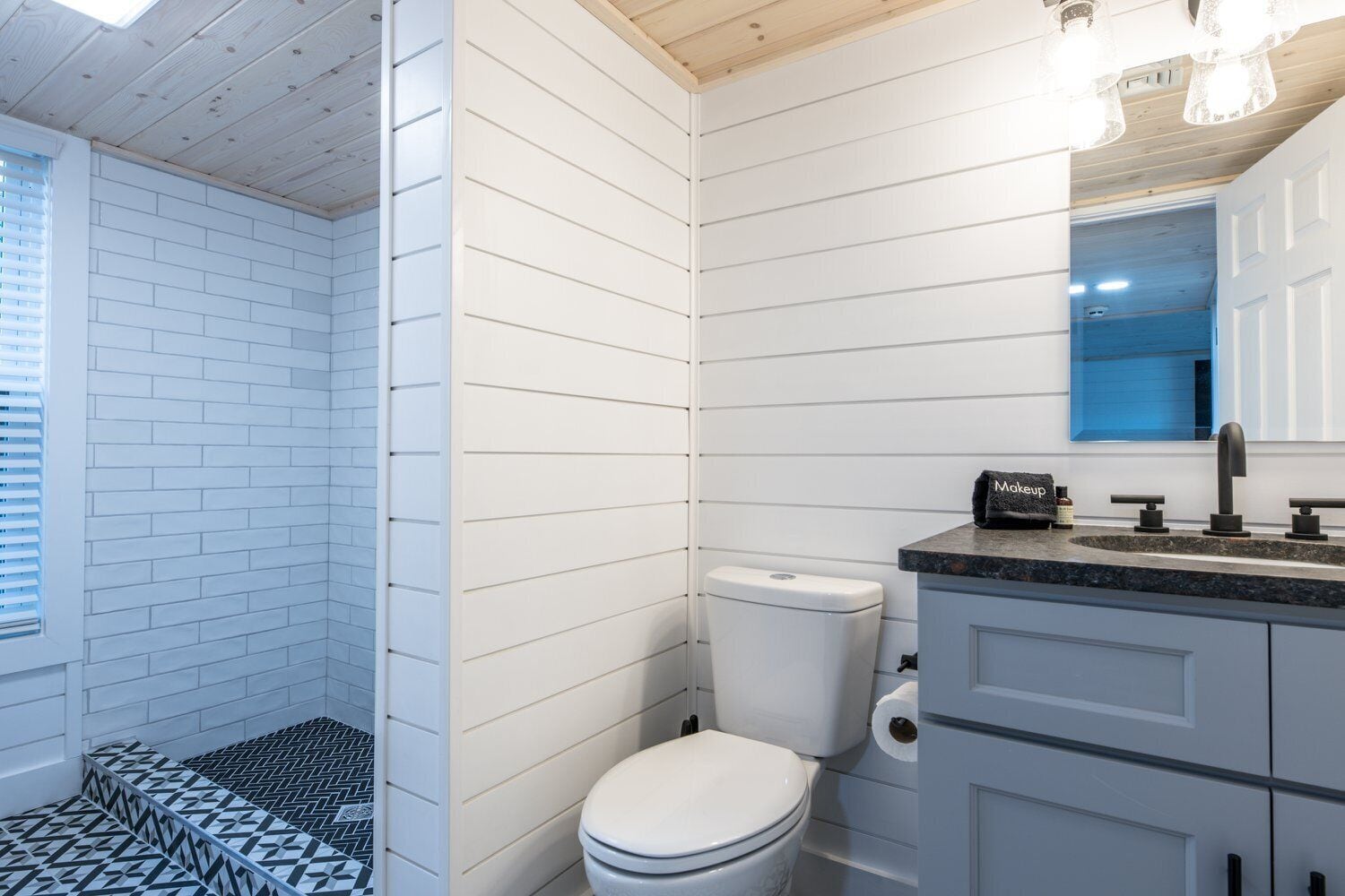 Un baño con paredes de azulejos blancos, inodoro blanco, piso de ducha con diseño blanco y negro y un lavamanos moderno con espejo arriba.