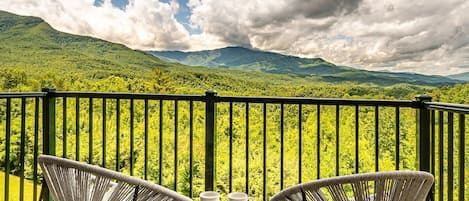 Ein Balkon mit zwei Korbstühlen und einem kleinen Tisch, mit Blick auf eine üppige, grüne Landschaft und Berge unter einem teils bewölkten Himmel.