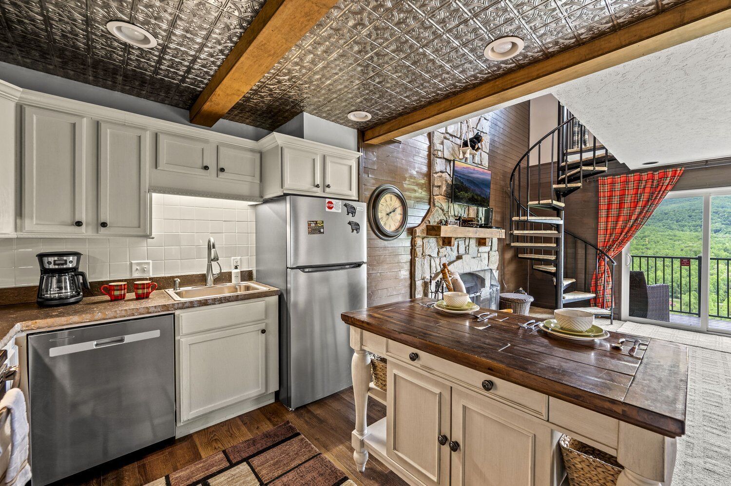A kitchen with white cabinets, a stainless steel refrigerator, and a wooden countertop. There is a stone fireplace and a staircase leading to a balcony with a red plaid curtain.
