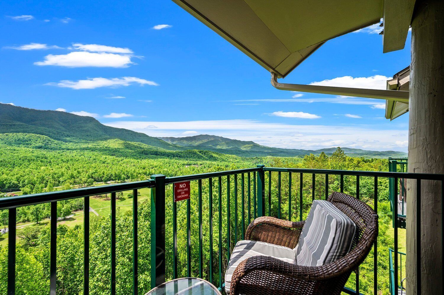 Un balcón con una silla de mimbre, vista a montañas verdes y un cartel que indica 'No Grilling or Smoking'.