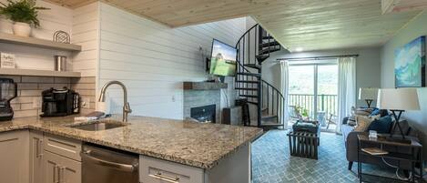 A modern kitchen with granite countertops, a built-in dishwasher, and a stainless steel sink. There is a spiral staircase leading to a balcony with a view of the outdoors.