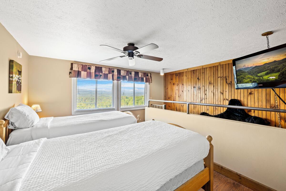 Ein Schlafzimmer mit zwei Betten, einem Deckenventilator, einem Fernseher und einem Fenster mit Blick auf Grünflächen.