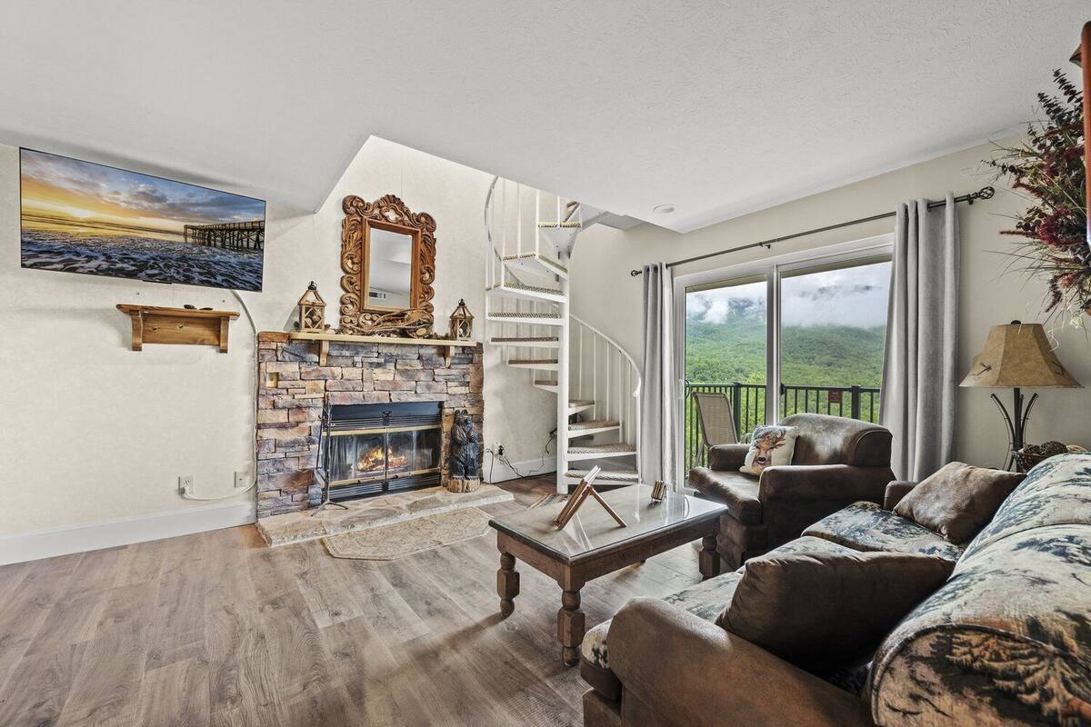 Ein Wohnzimmer mit Kamin aus Stein, einem Couchtisch aus Holz, einer Couch und einer Treppe zu einem Balkon mit Blick ins Grüne.