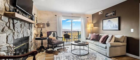A living room with a stone fireplace, a sofa, a coffee table, and a view of the outdoors.
