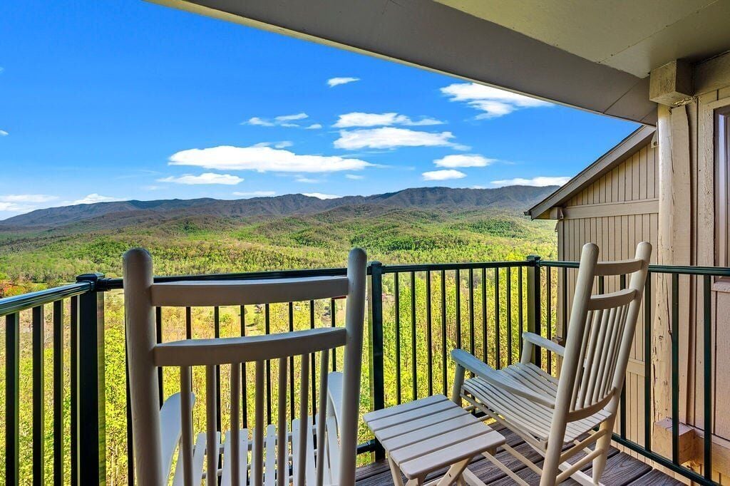 A balcony with two white Adirondack chairs and a view of green hills and mountains.