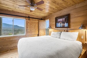 A bedroom with a large bed, wooden walls, a ceiling fan, a window with a mountain view, and a framed picture on the wall.