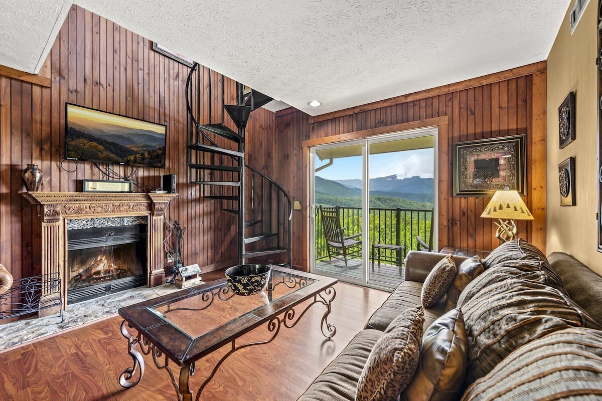 Ein gemütliches Wohnzimmer mit Kamin, einer Holztreppe und einem Balkon mit Bergblick.