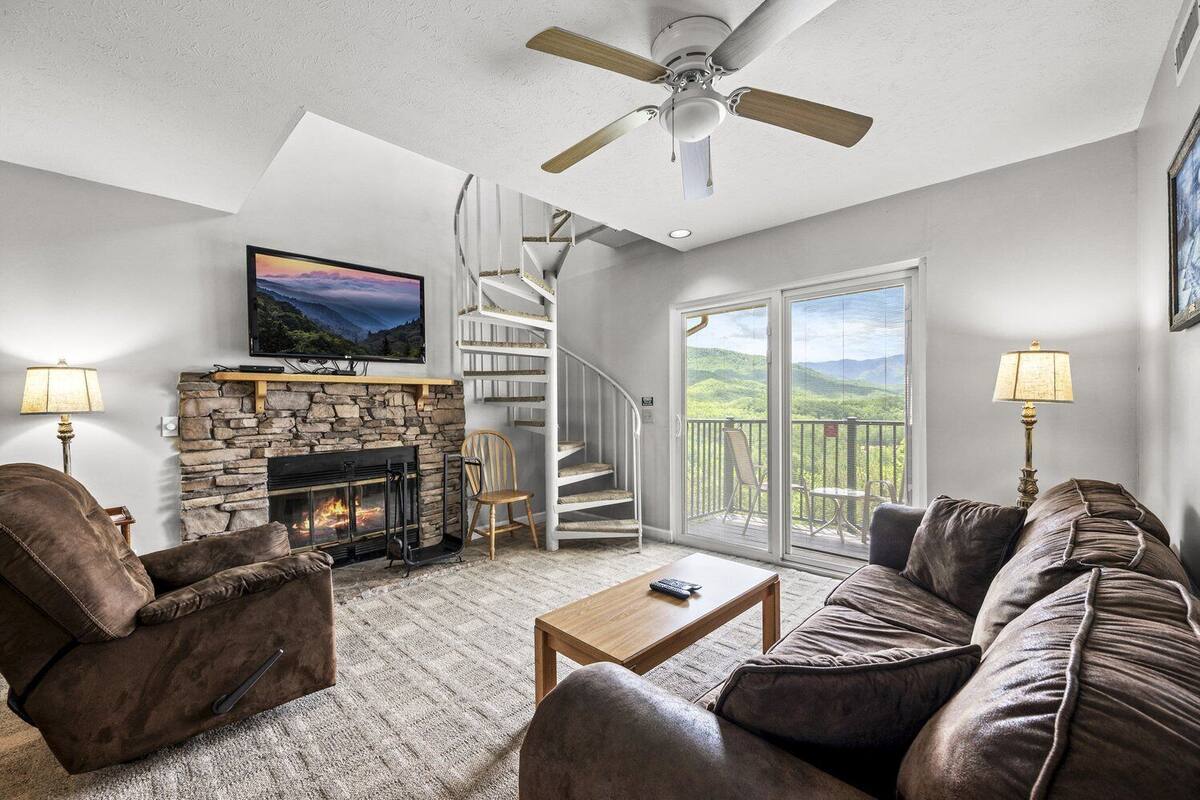 Ein Wohnzimmer mit Kamin aus Stein, einer Couch, einem Couchtisch und einem Fernseher über dem Kamin. Es gibt einen Deckenventilator, eine Treppe mit einer Glastür, die zu einem Balkon führt, und zwei Lampen auf beiden Seiten der Couch.