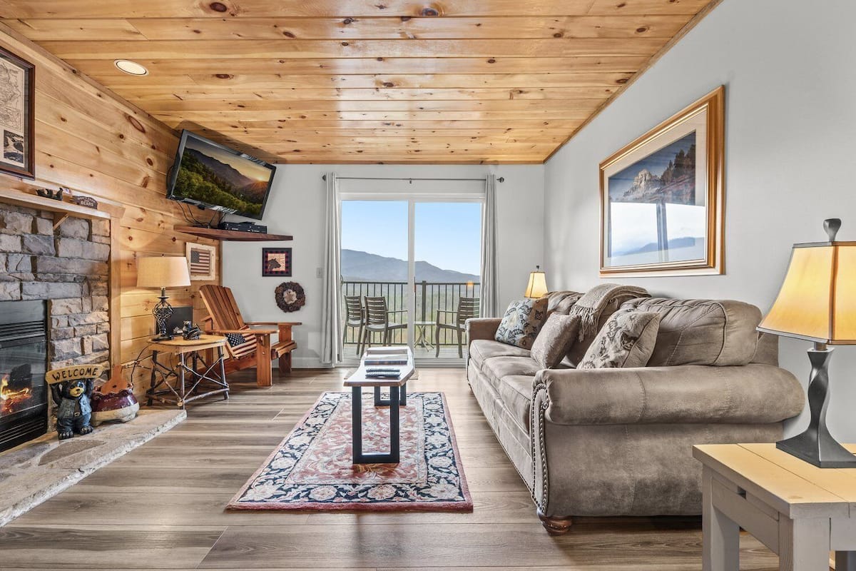 Ein gemütliches Blockhauss-Interieur mit Kamin, Holzbalkendecke, einer Couch und einem Balkon mit Blick.
