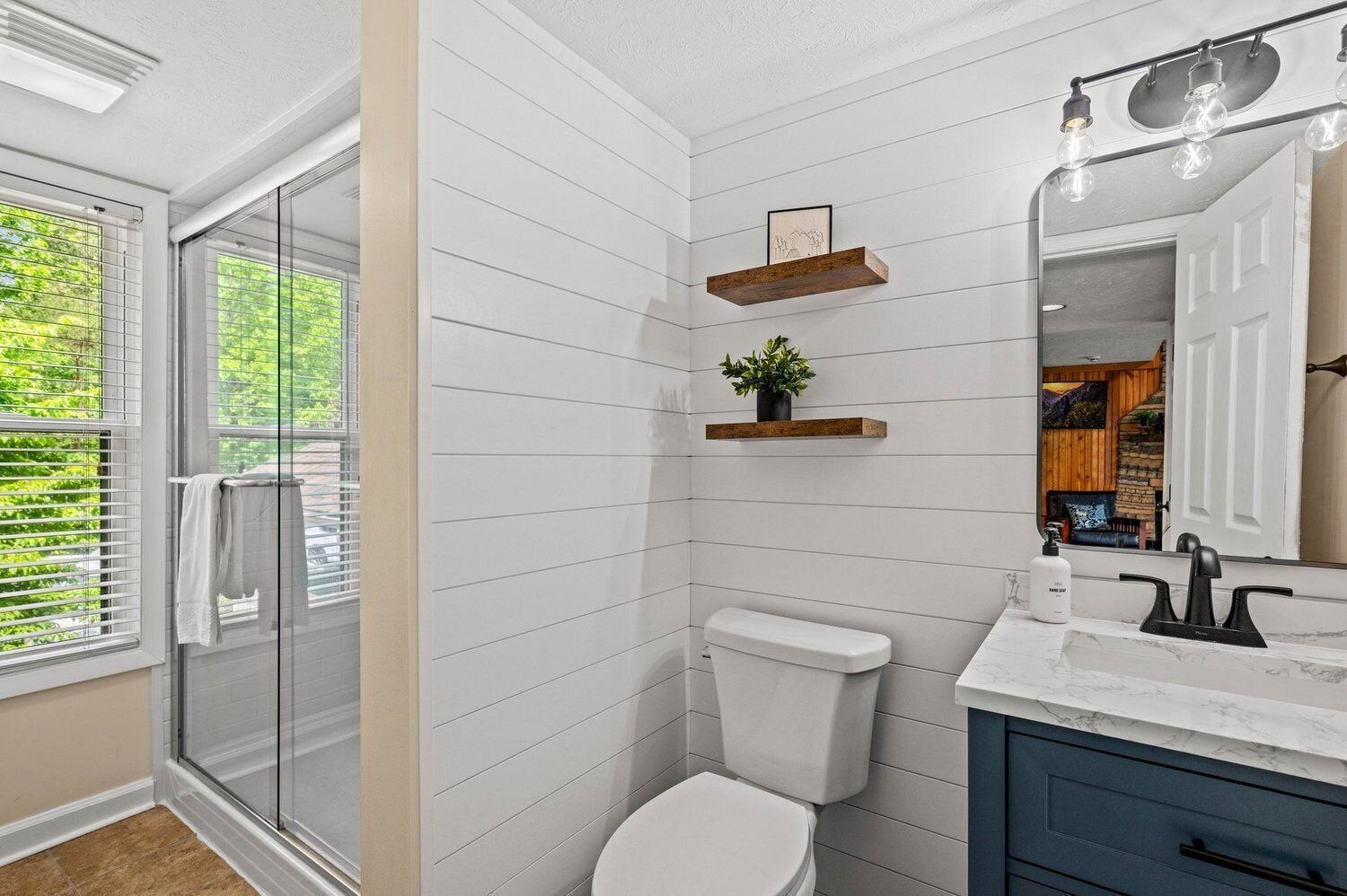 A bathroom with a white tiled wall, a white toilet, a wooden shelf with a plant, a marble countertop, and a large mirror.