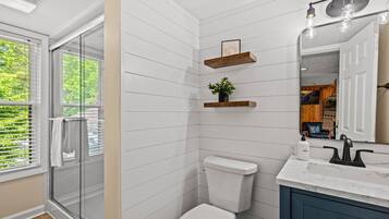 A bathroom with a white tiled wall, a white toilet, a wooden shelf with a plant, a marble countertop, and a large mirror.