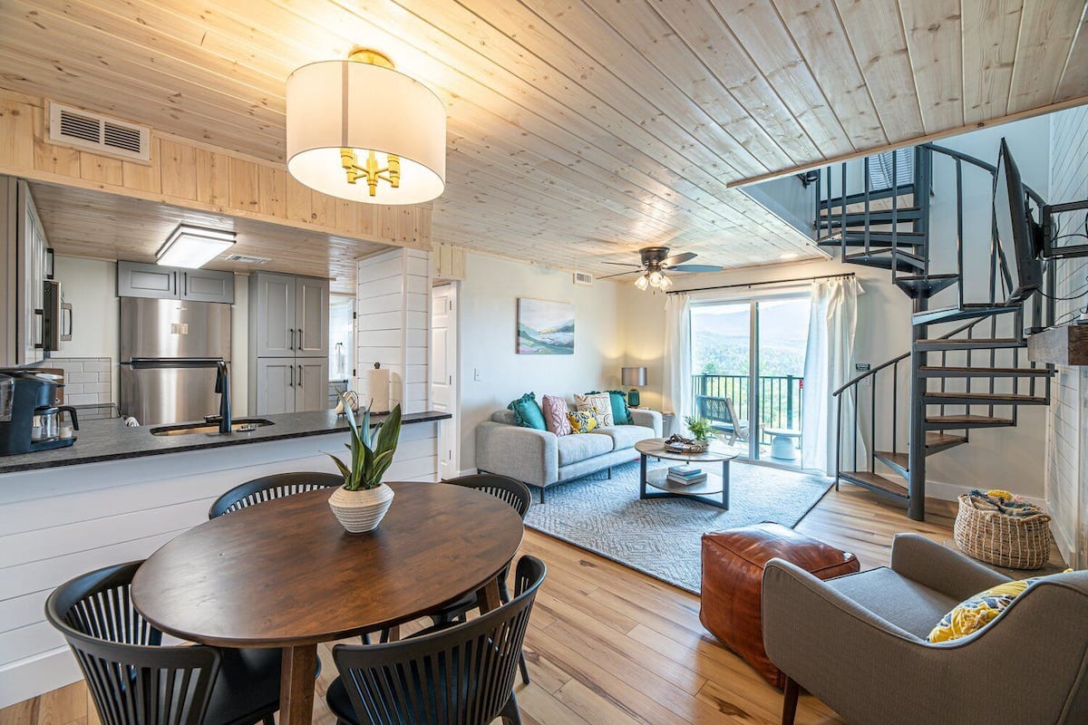 Eine moderne Küche mit Edelstahlgeräten, ein Essbereich mit Holztisch und -stühlen, ein Wohnzimmer mit Sofa und Couchtisch sowie eine Wendeltreppe, die zu einem Balkon führt.