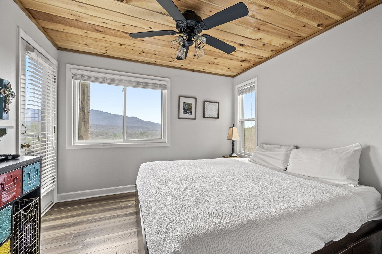 Un dormitorio con techo de madera, una cama con ropa de cama blanca, dos ventanas y un ventilador de techo.