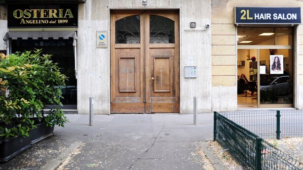 Un edifici amb una porta doble de fusta, un rètol que diu 'Osteria Angelino dal 1899' i un rètol que anuncia '2L Hair Salon'.