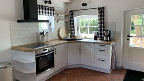 A compact kitchen with white cabinets, a built-in oven, a coffee maker, and a window with checkered curtains. - Hygge-huus-burhave Comfortable Holiday Residence (Butjadingen)
