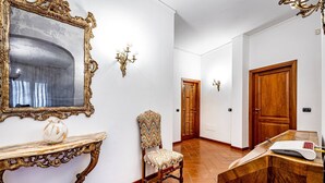 Een gang met een grote, versierde spiegel, een houten stoel, een console tafel met een vaas en een houten deur.