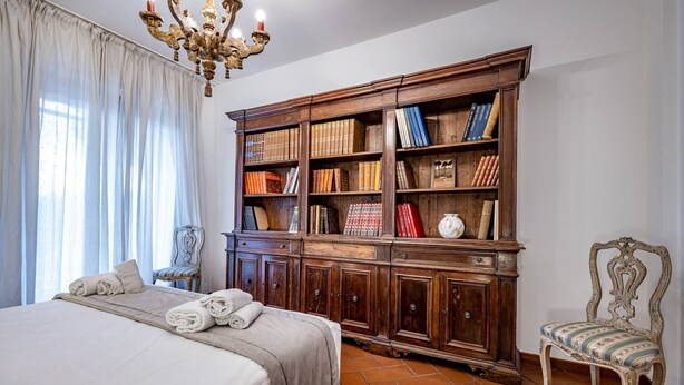 Een slaapkamer met een groot bed, een houten boekenkast, een kroonluchter en een vintage fauteuil.