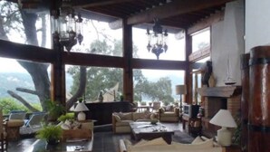 A spacious living room with a fireplace, comfortable seating, and large windows offering a view of the outdoors. - Casa Vallesana, Fraccionamiento "El Coporito", Magnificent lake view (Valle de Bravo)