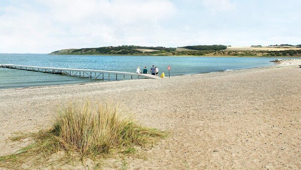 A beach with a pier extending into calm waters, a grassy area in the foreground, and a hilly landscape in the background. - 5 Person Holiday Home on a Holiday Park in Lemvig (Lemvig)
