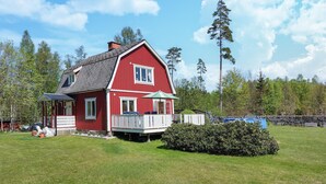 Une maison rouge avec un porche et une petite terrasse, entourée d’une pelouse bien entretenue et d’arbres.
