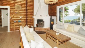 Ein gemütliches Blockhaus-Interieur mit Kamin, weißem Sofa, Couchtisch aus Holz und einem großen Fenster mit Blick auf eine reizvolle Landschaft.
