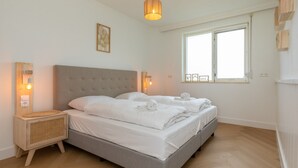 A modern bedroom with a large bed, bedside lamps, a wooden nightstand, and a window with a view. - Charming Holiday Home With a Terrace (Zoutelande)