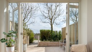An open patio with a view of a garden, a white table with a candle, and a beige sofa. - Charming Holiday Home With a Terrace (Zoutelande)