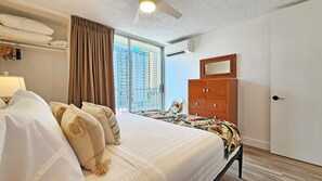 Une chambre d’hôtel avec un lit, une armoire en bois, une fenêtre donnant sur des immeubles de grande hauteur et un ventilateur de plafond.