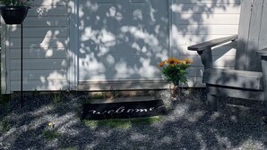 Une maison en bois blanche, avec une porte d’entrée, un paillasson, un banc et un pot de fleurs.