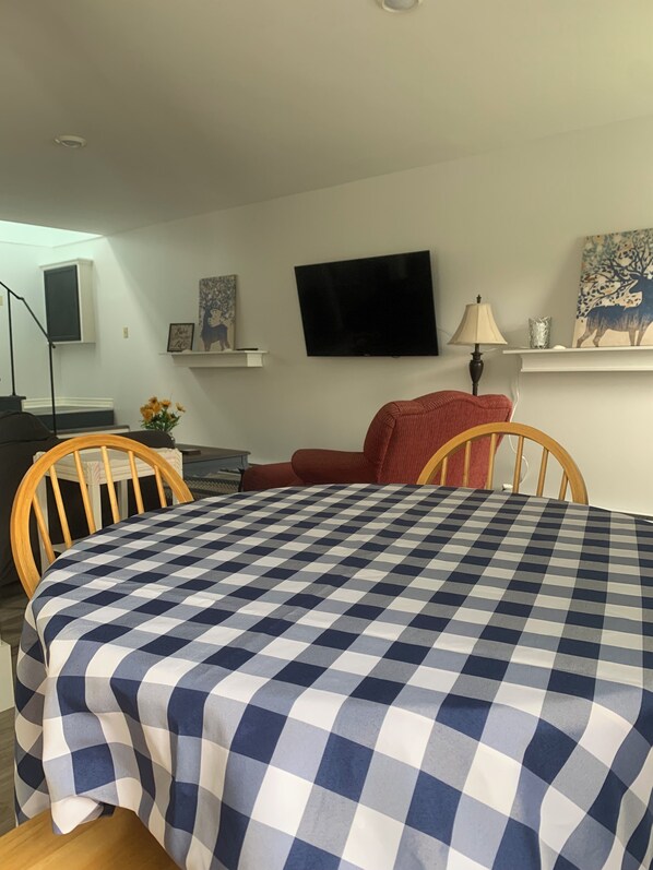 Ein Essbereich mit kariertem Tischläufer, Holzstühlen, einem Fernseher an der Wand und einer Lampe auf einem Beistelltisch.