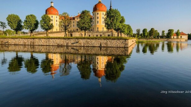 Ein historisches Gebäude mit zwei markanten Kuppeln, einer Steinmauer und einem ruhigen Gewässer, das die Struktur reflektiert.
