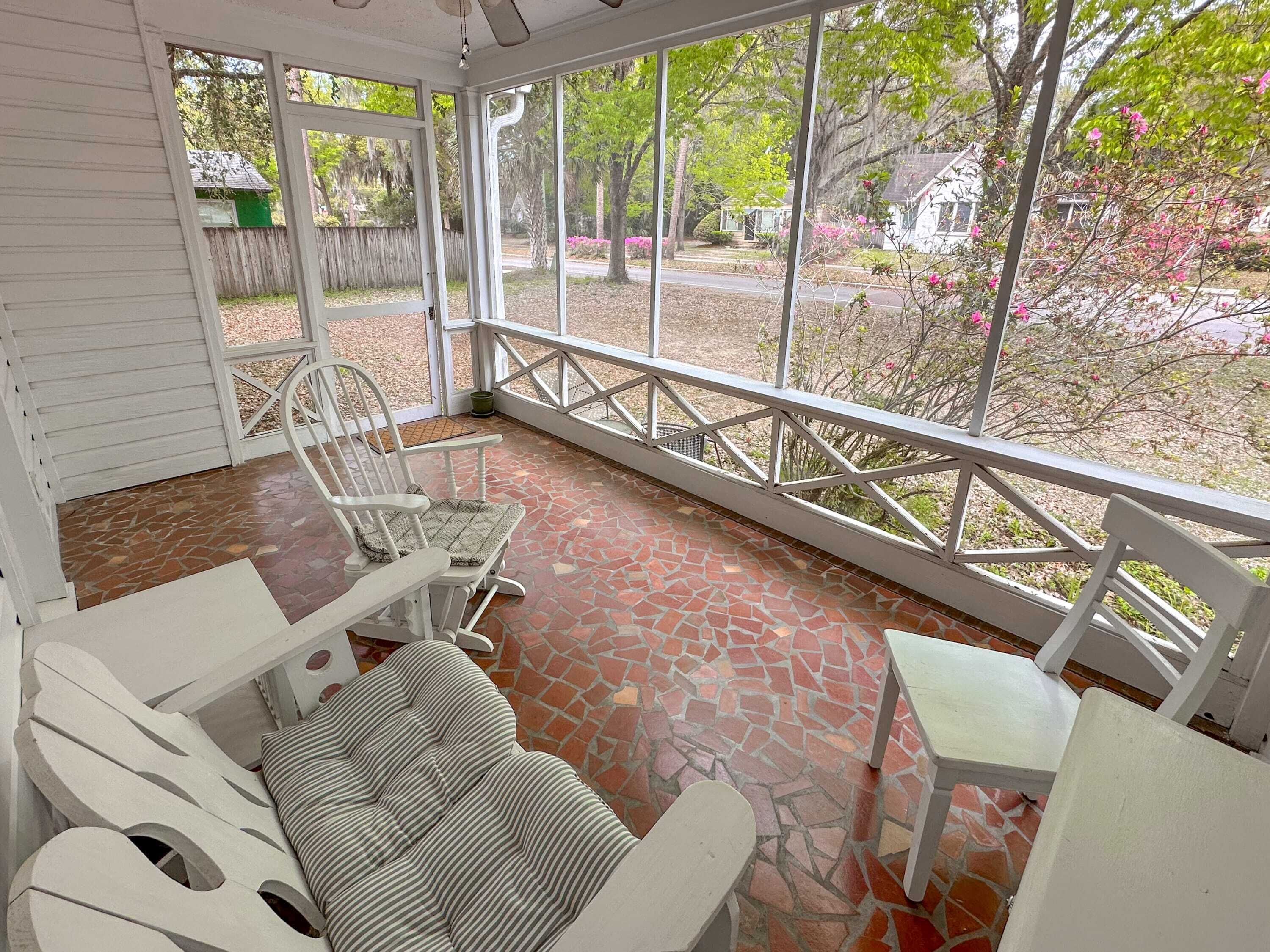 Une terrasse couverte avec un sol carrelé, des meubles blancs et de grandes fenêtres donnant sur un quartier résidentiel avec des arbres et des maisons.