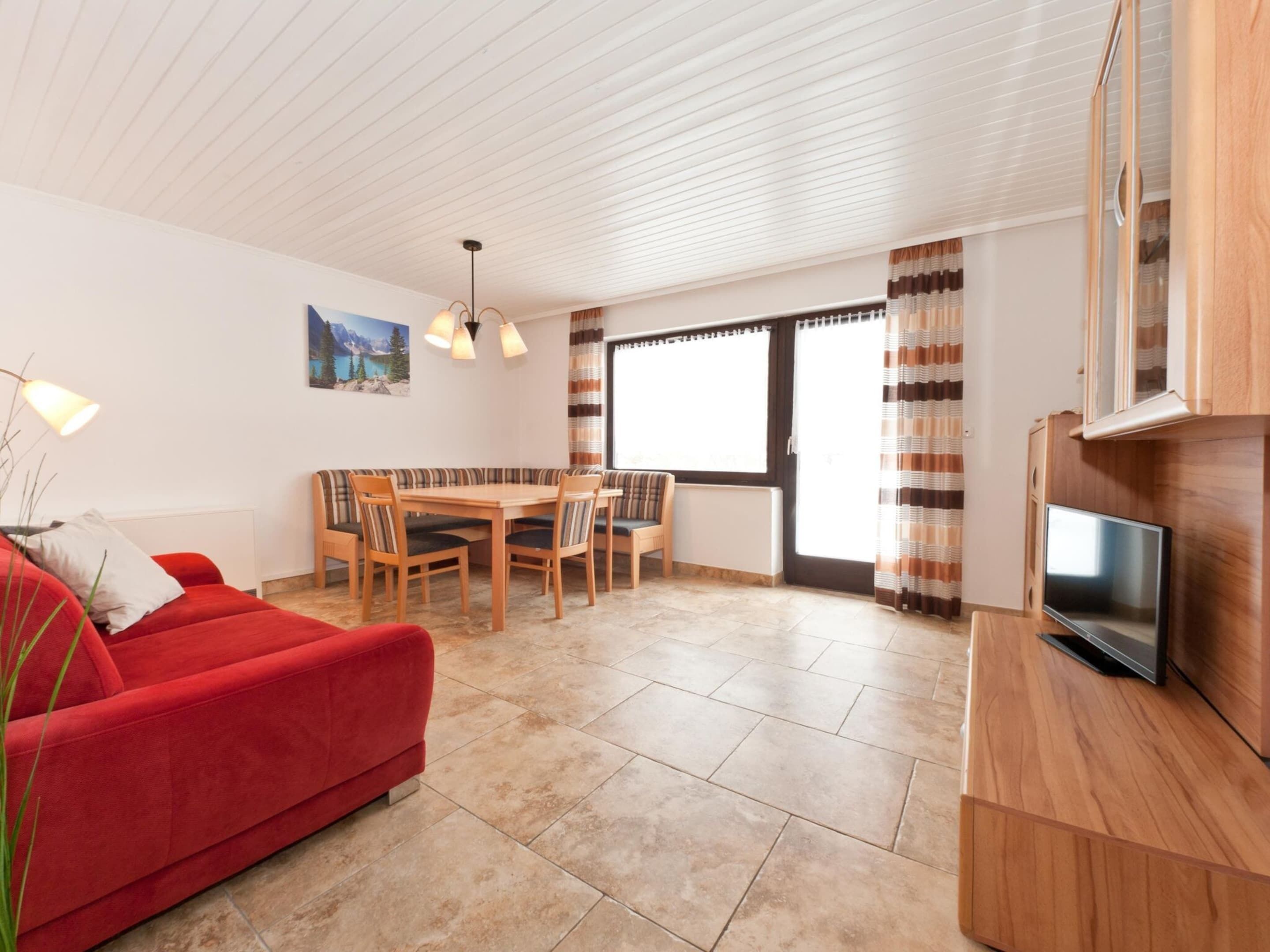 Un salon moderne avec un canapé rouge, une table à manger avec des chaises, un téléviseur posé sur un meuble en bois et une grande fenêtre avec des rideaux.