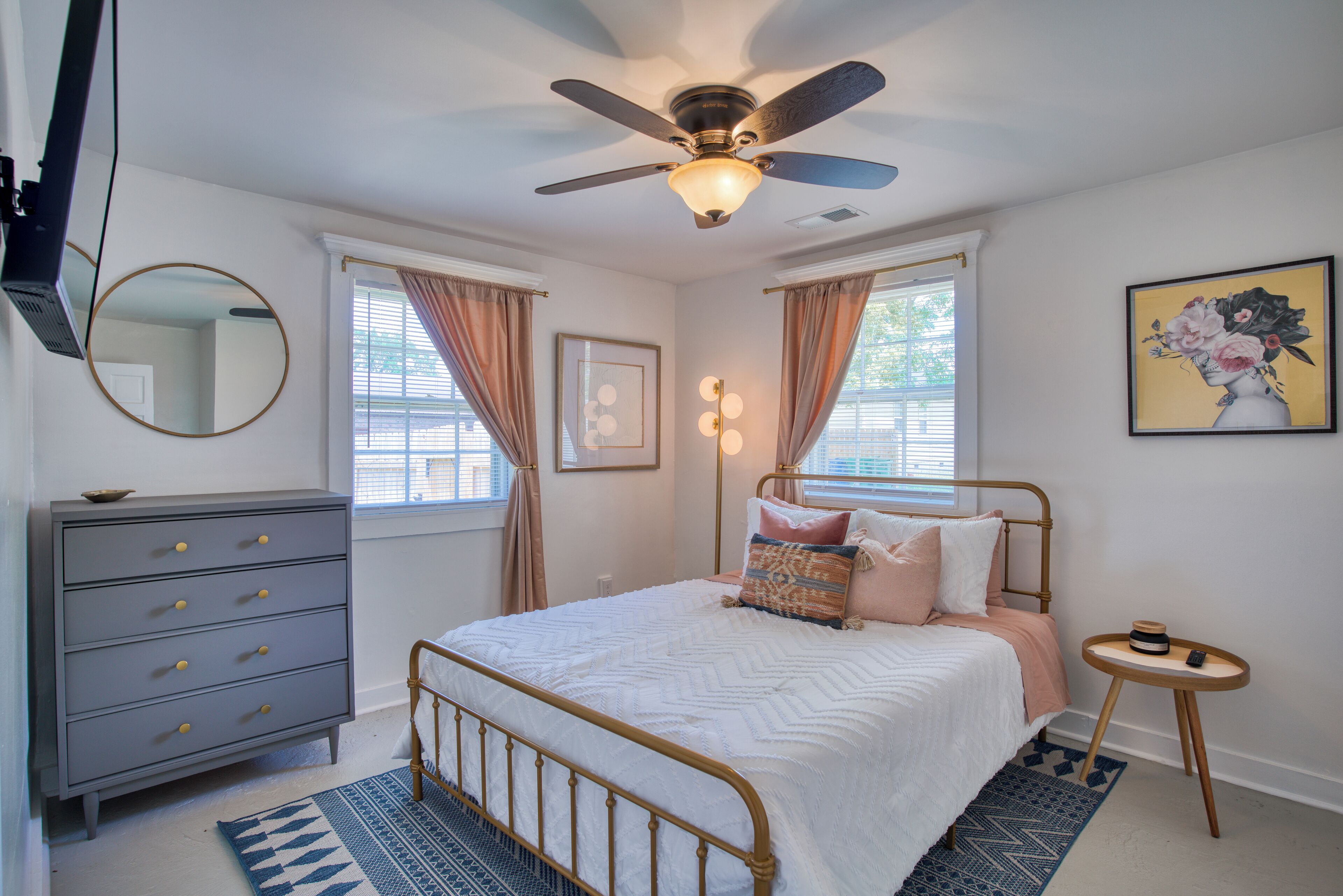 Une chambre à coucher comprenant un lit, une commode, un ventilateur de plafond, des fenêtres et des œuvres d’art.