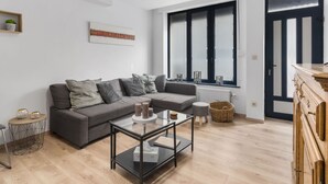 A modern living room with a grey sofa, a glass coffee table, and a large window with blinds. - Beachfront Holiday Home Near the Market (Blankenberge)