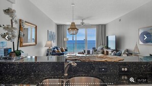 A modern living room with a large window overlooking the sea, a central island with a sink, and a ceiling fan. - Beach Lovers Dream- 2 BR Majestic Sun Condo Sleeps 8! (Destin)