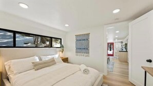 A bedroom with a large bed, a nightstand, a wall-mounted artwork, and a view into the adjacent kitchen.