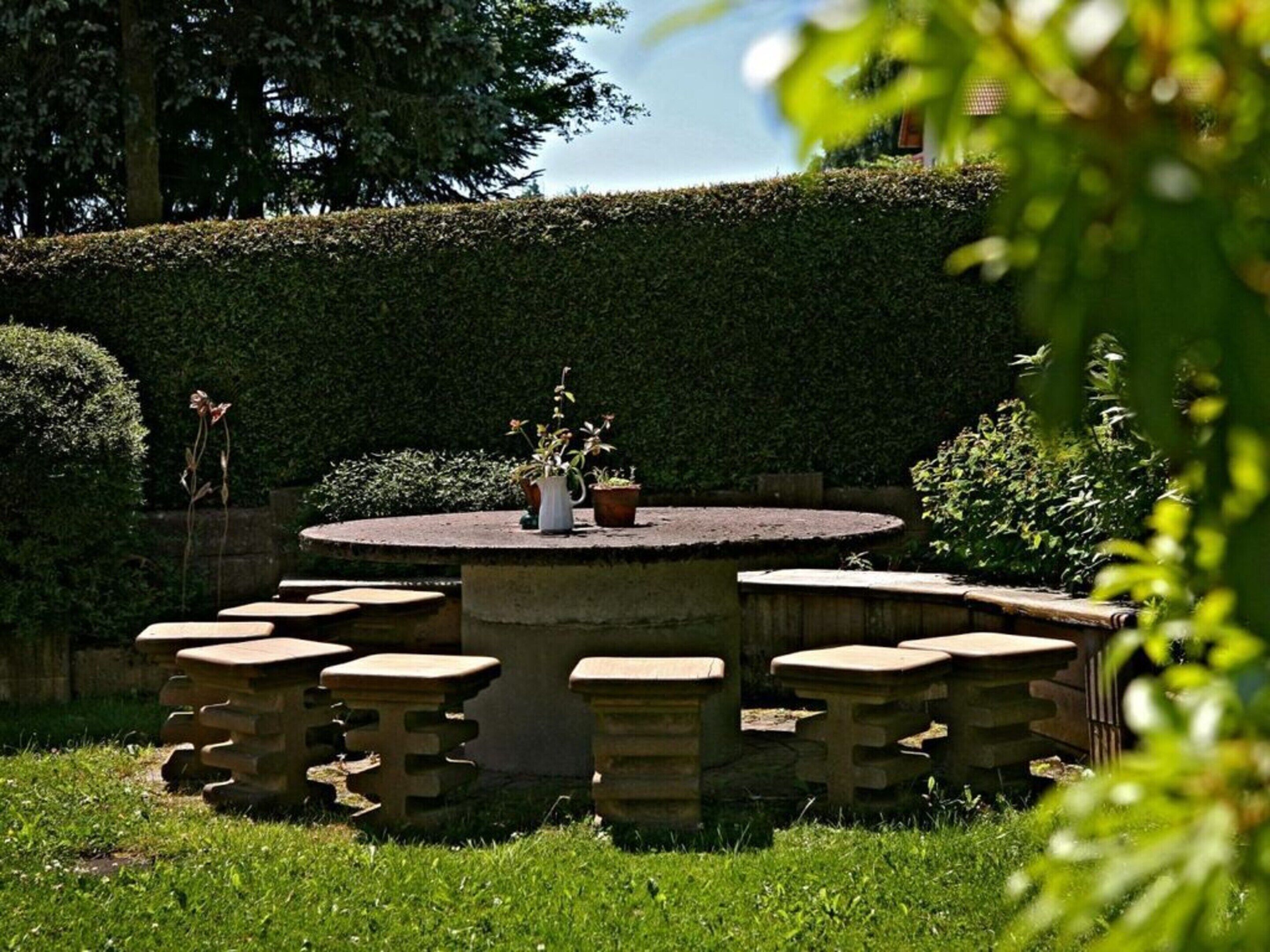A circular stone table with six stools, surrounded by a well-maintained hedge.