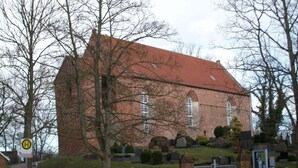 Un grand bâtiment en briques rouges, doté d’un toit en pente, vraisemblablement une église ou une chapelle, entouré d’un cimetière avec des pierres tombales et une entrée fermée par un portail.