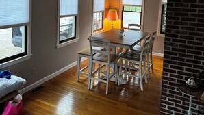A dining area with a wooden table and chairs, large windows, and a brick accent wall. - Last minute Cape cod rental
  Bikes,  Beach ,ac  ,laundry ,ping pong table  (Dennis)
