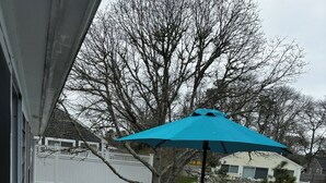 A wooden deck with a table and chairs, a blue umbrella, and a white railing. - Last minute Cape cod rental
  Bikes,  Beach ,ac  ,laundry ,ping pong table  (Dennis)