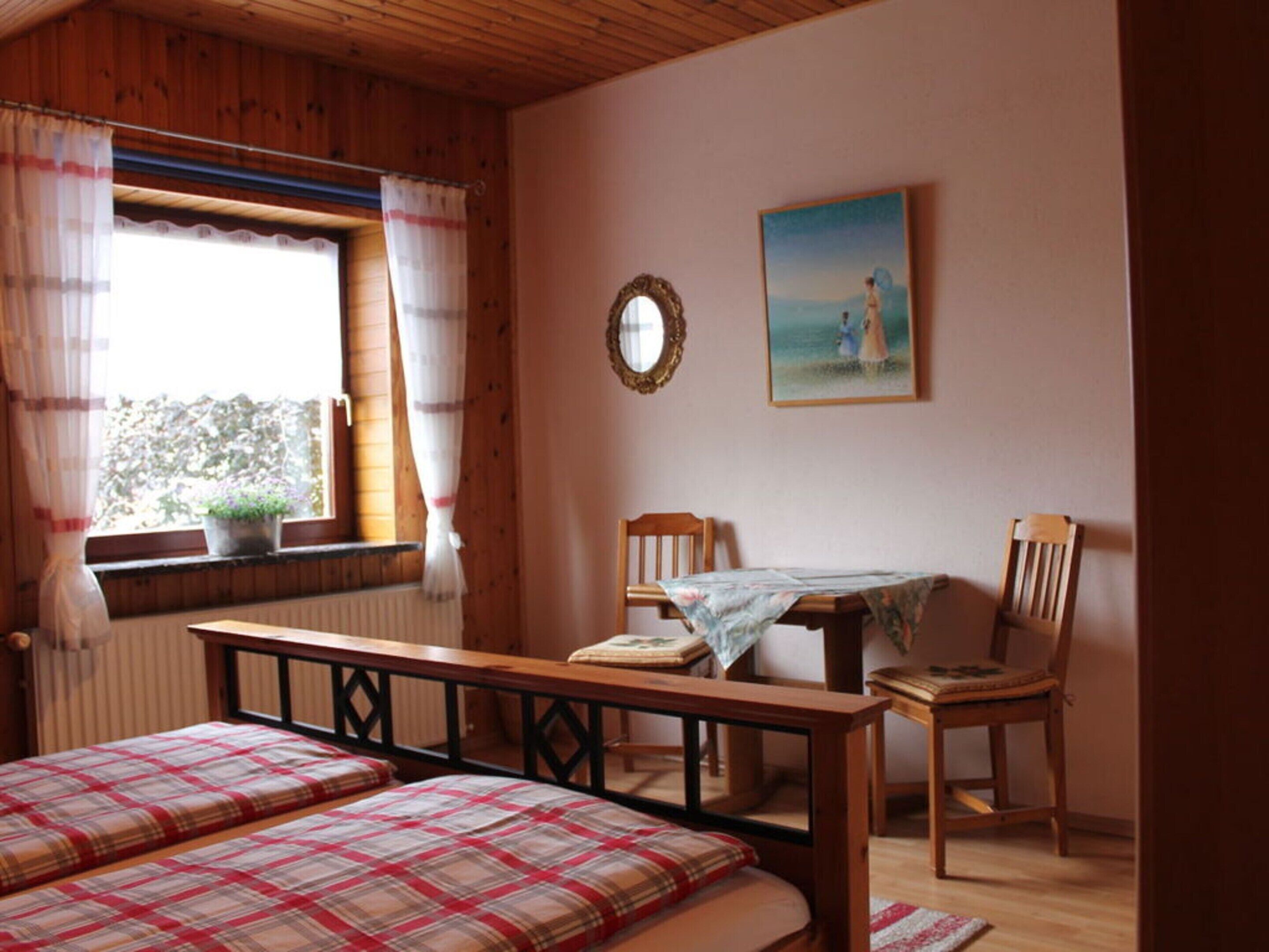 A cozy room with a bed, a table, and two chairs. There's a window with curtains and a painting on the wall.